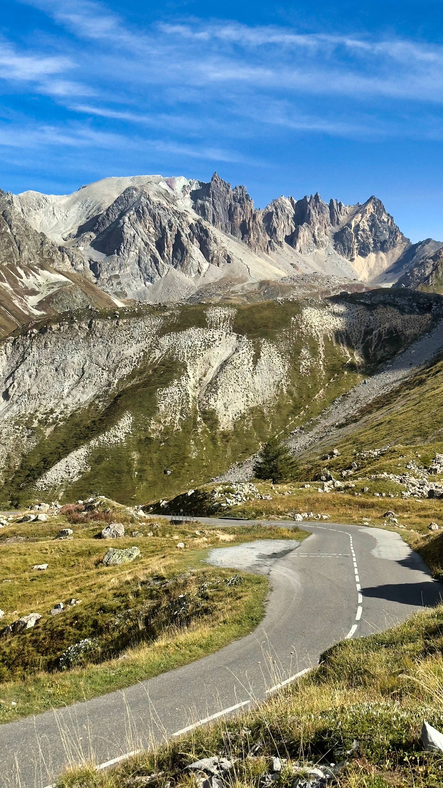 A narrow paved mountain road curving through alpine terrain with jagged granite spires in the distance