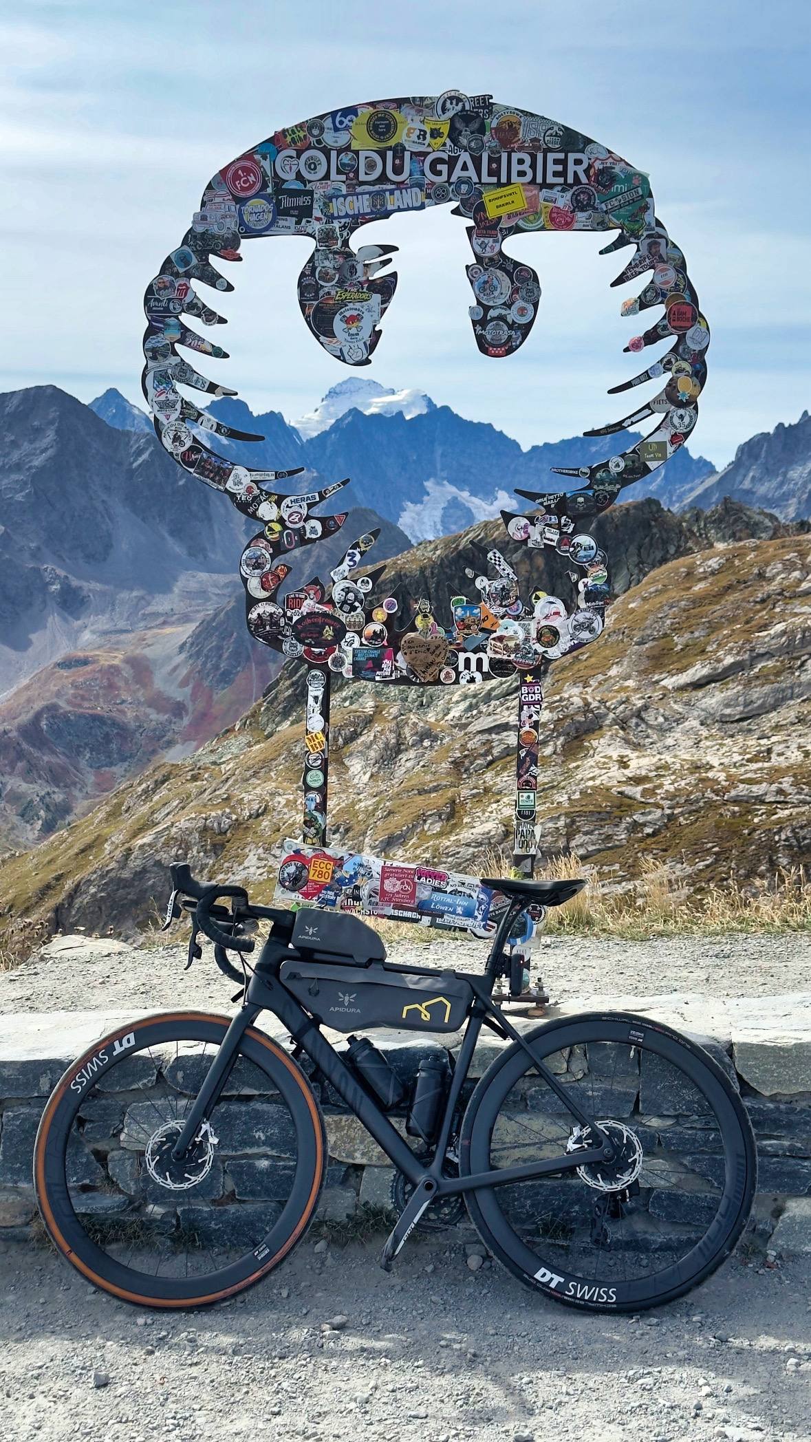 A gravel road bike loaded with bikepacking bags leaning against the Col du Galibier summit sign, jagged peaks behind