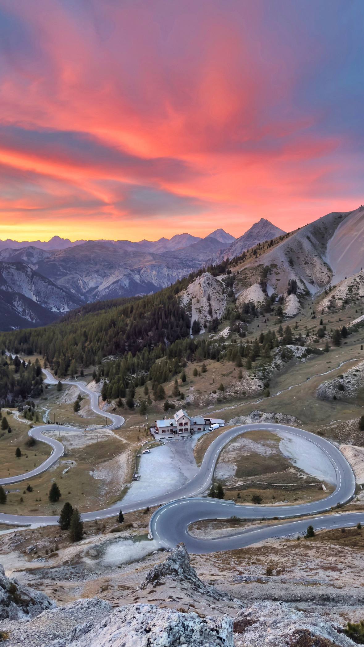 A dramatic pink and orange sunset over hairpin switchbacks and a mountain refuge, with peaks silhouetted on the horizon
