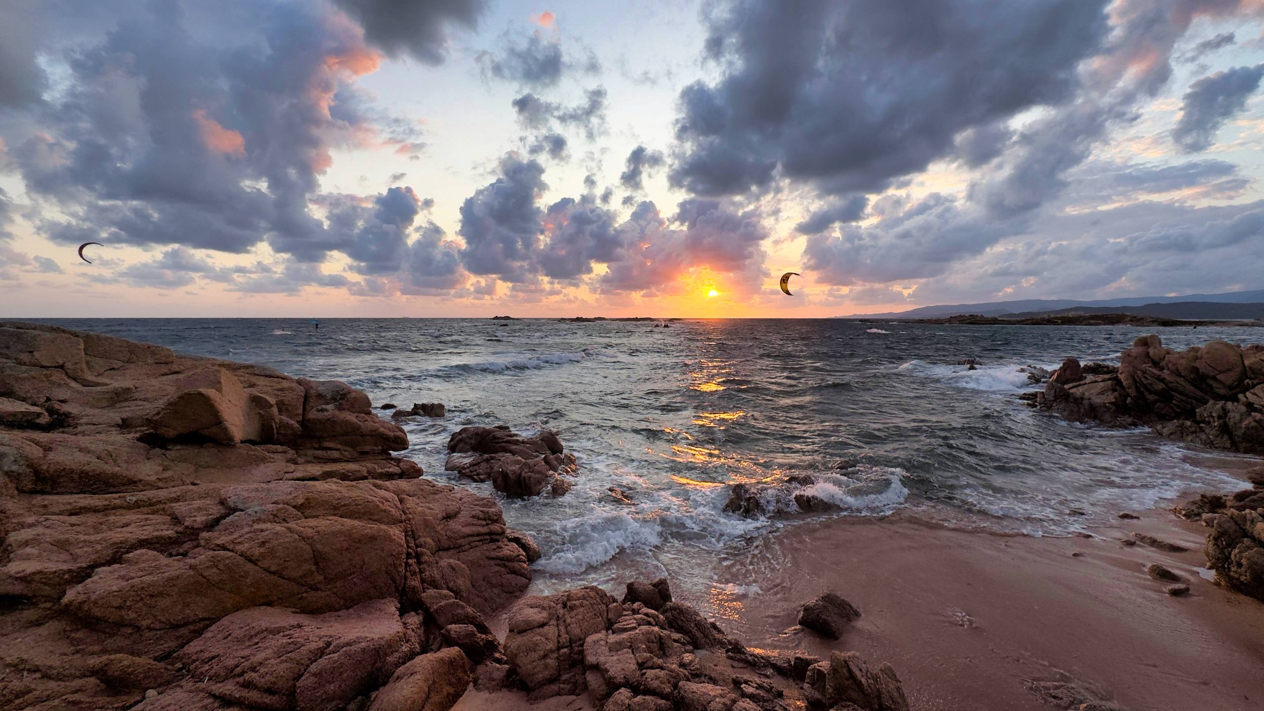 Sunset over the sea with kitesurfers silhouetted and waves on pink granite rocks