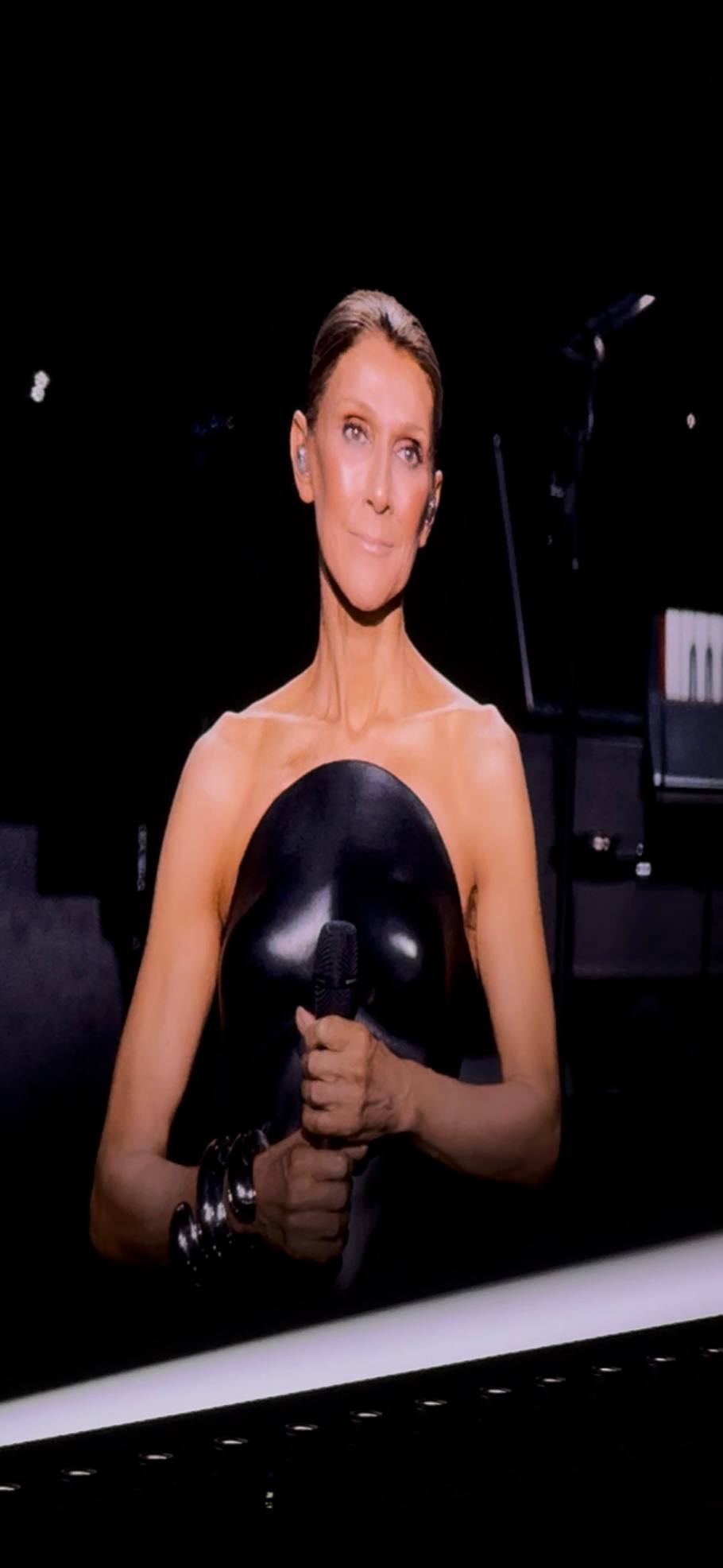 Céline Dion performing on stage in a black gown, close-up on a large arena screen