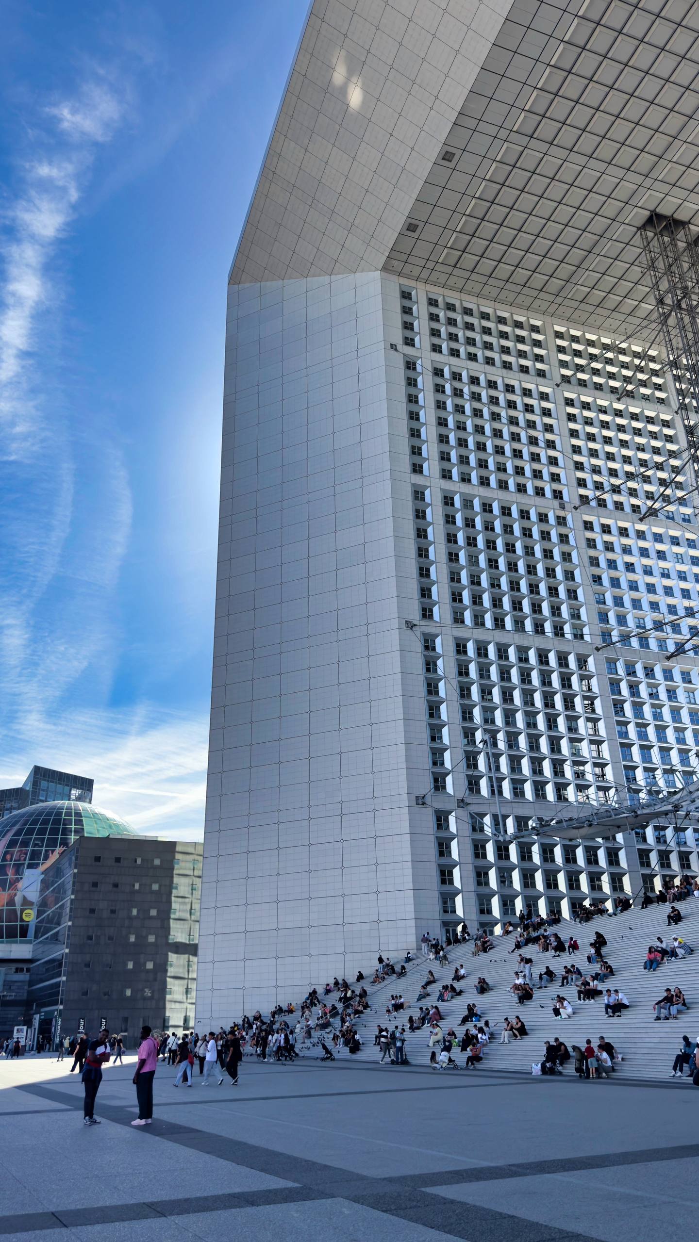 Looking straight up at the underside of the Grande Arche de la Défense, a massive white cube-shaped monument, with crowds sitting on the steps below