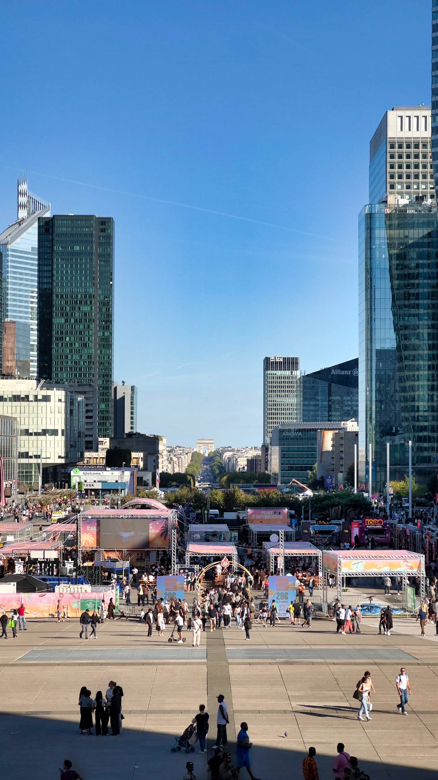 The skyline of La Défense business district with skyscrapers framing a view down the axis toward the Arc de Triomphe, a public square and fair in the foreground