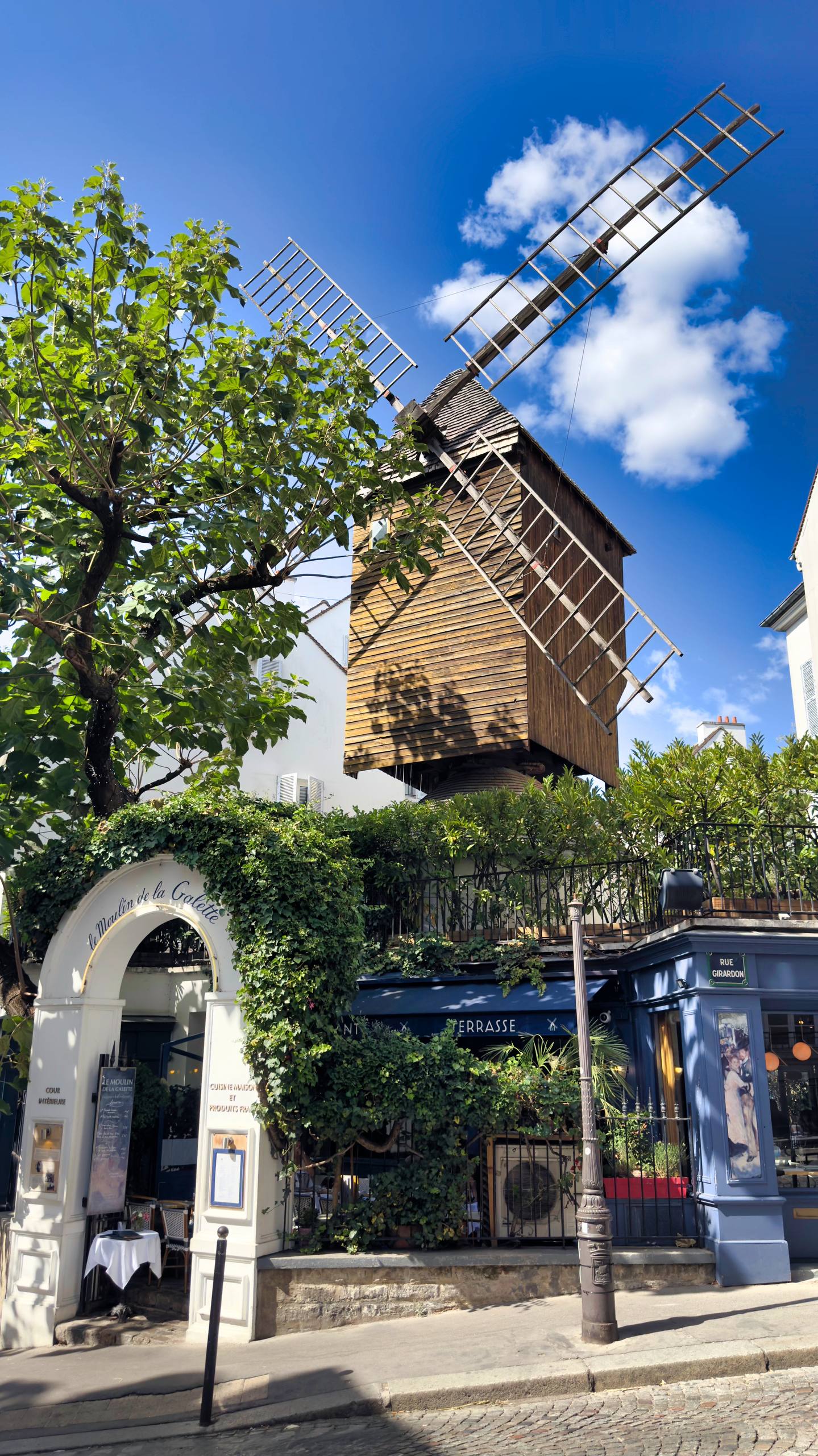 The wooden windmill of Le Moulin de la Galette in Montmartre, framed by a chestnut tree against a blue sky