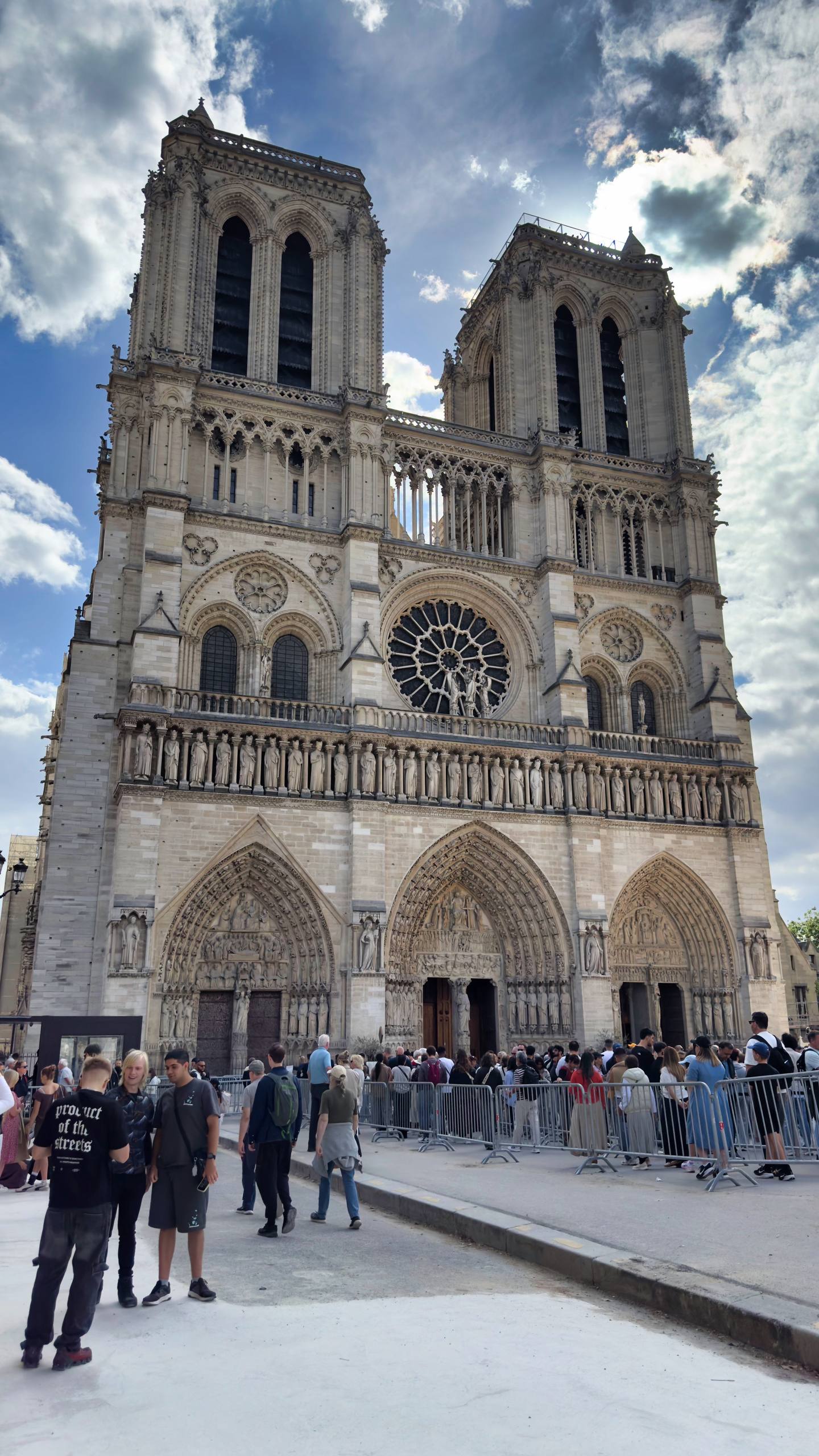 The twin Gothic towers and rose window facade of Notre-Dame cathedral, with crowds queuing behind barriers on the square below
