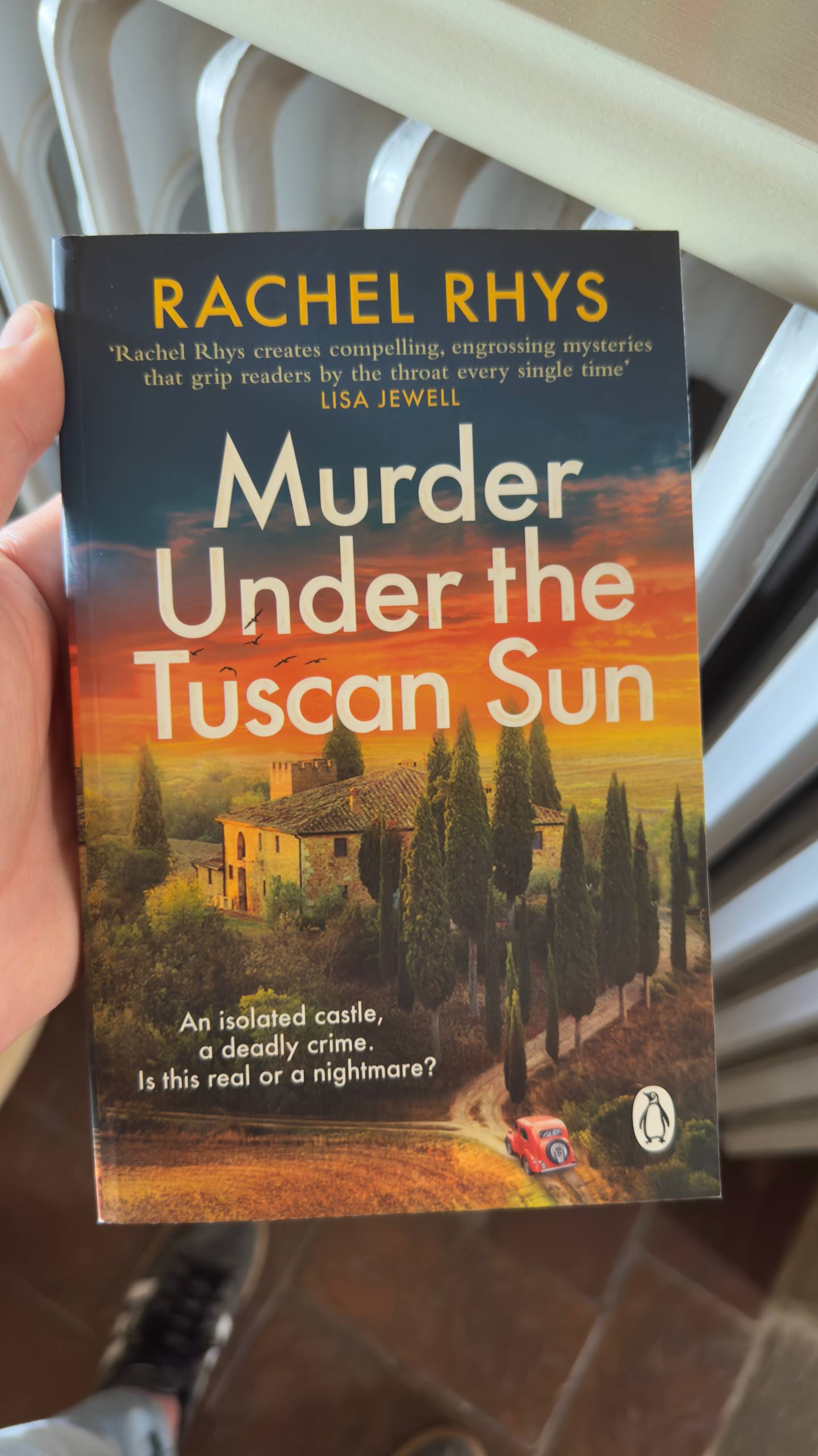 A hand holding the paperback Murder Under the Tuscan Sun by Rachel Rhys, with a sunset Tuscan villa on the cover