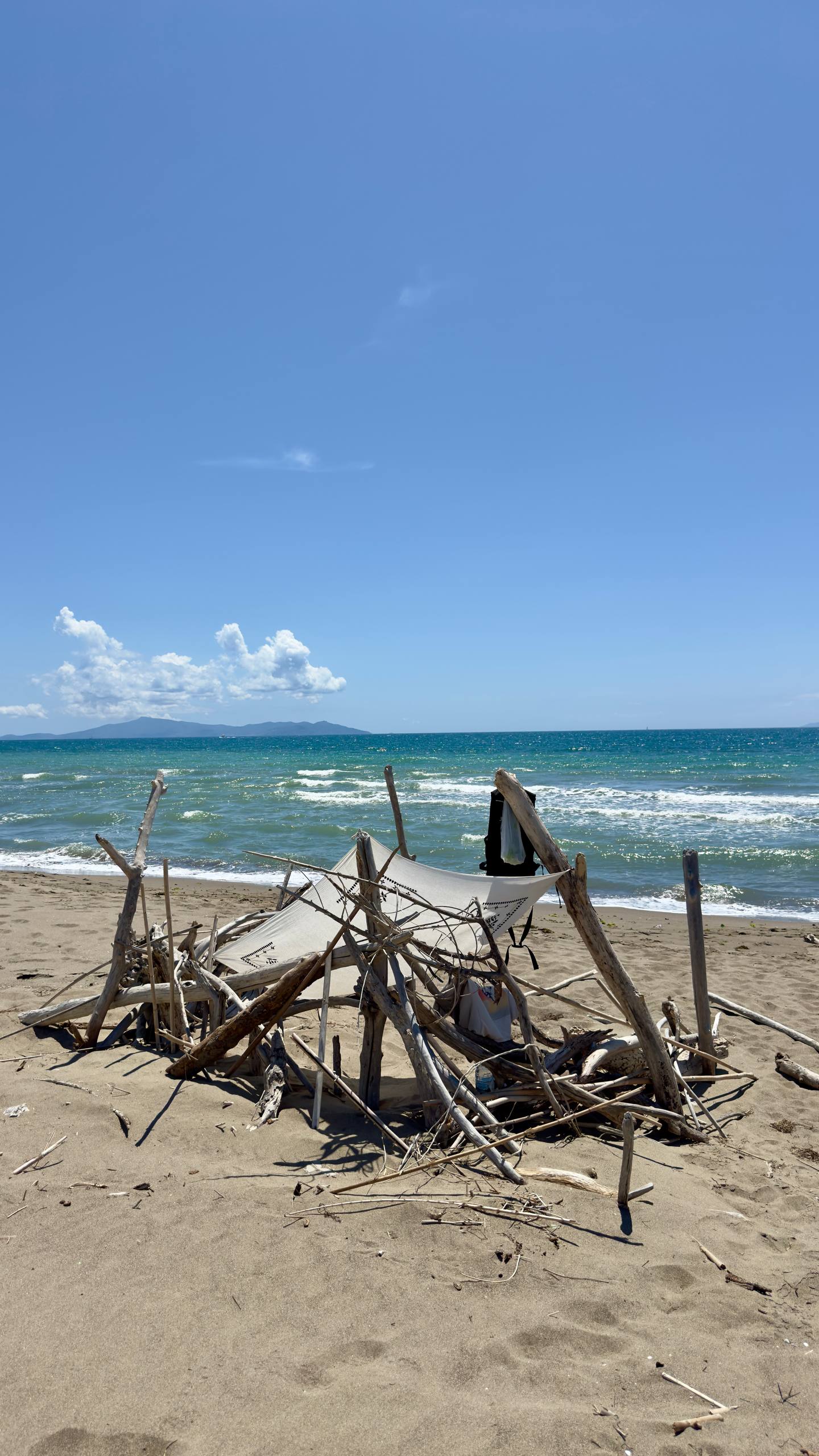 A driftwood teepee shelter on an empty sandy beach with turquoise Mediterranean waves and an island silhouetted on the horizon