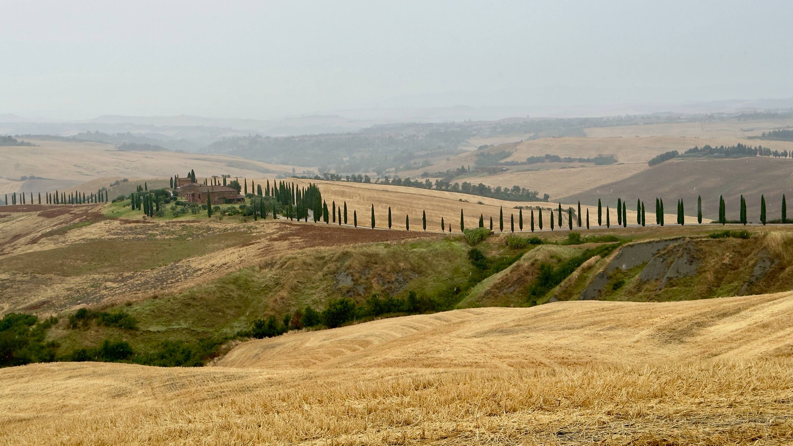A stone farmhouse on a hilltop with a cypress-lined driveway through harvested wheat fields in the Val d'Orcia