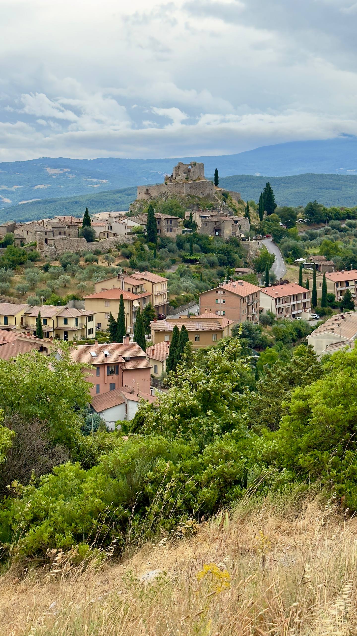 A Tuscan hilltop village crowned by a ruined medieval castle, with terracotta roofs and olive groves cascading down the slope
