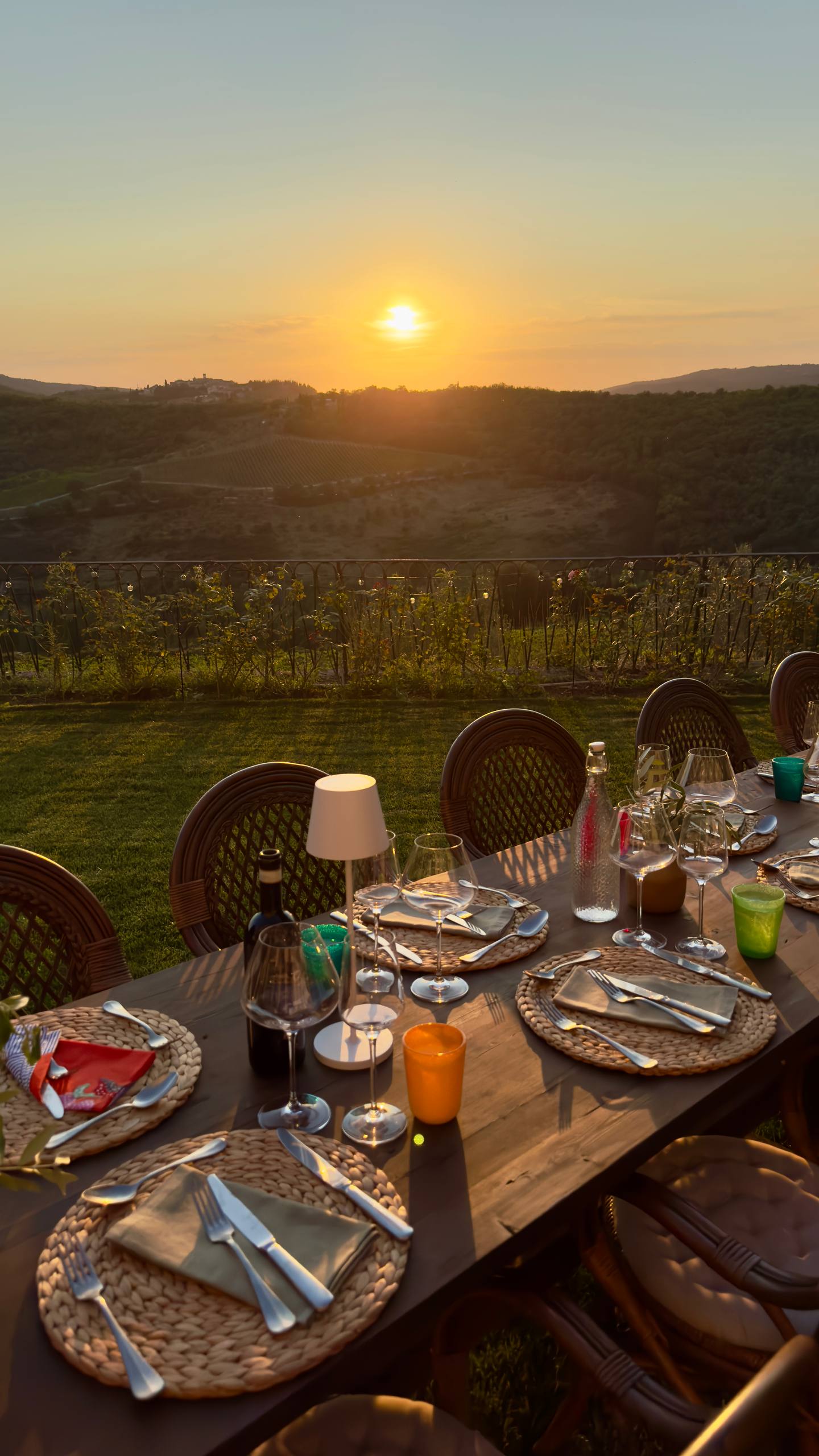 A long outdoor dinner table with woven placemats, wine glasses and candles, overlooking Tuscan vineyards at golden hour